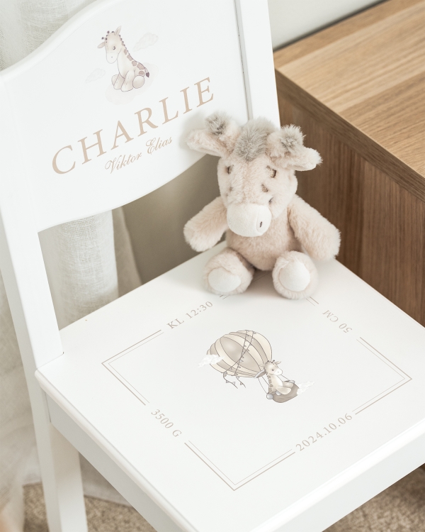 Vit dopstol med namn på ryggstödet, samt datum, tid och giraffmotiv på sitsen