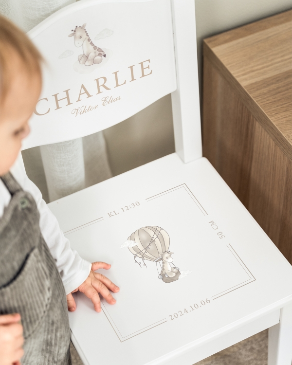 Vit dopstol med namn på ryggstödet, samt datum, tid och giraffmotiv på sitsen